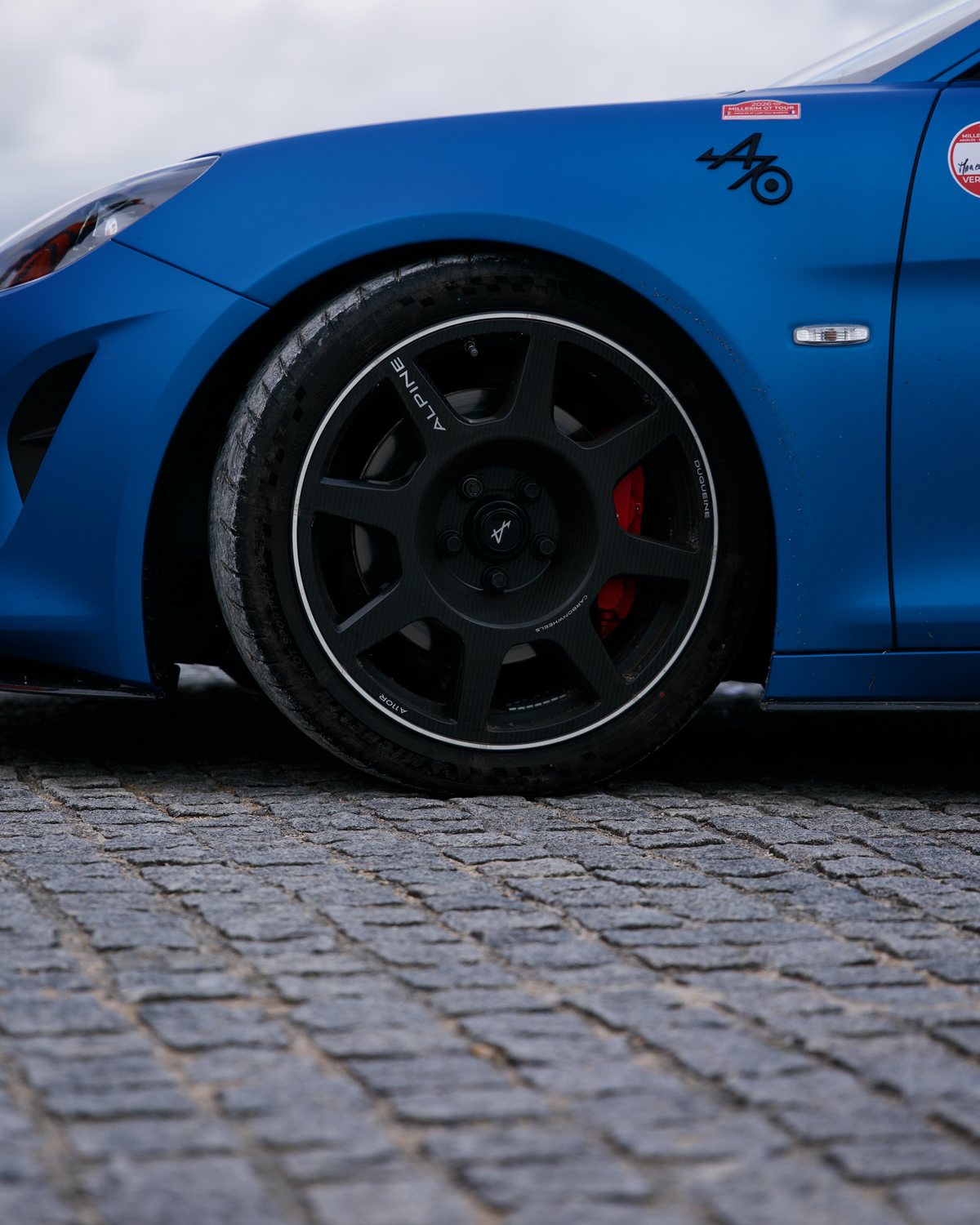 Roue en carbone et aile bleu mat d'une Alpine A110 posée sur des pavés, avec un autocollant Millesim GT Tour