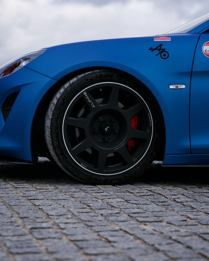 Roue noire d'une Alpine A110 bleu mat posée sur des pavés